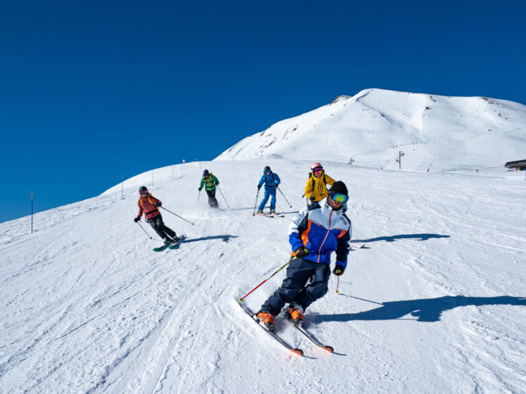 En gruppe skiløbere iført farverige tøj går ned ad en snedækket skråning på en solskinsdag. Himlen er klar, og en sneklædt bjergtop er synlig i baggrunden. De står på ski i en V-formation og skaber en dynamisk actionscene.