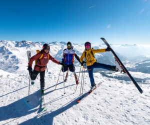 Tre skiløbere i farverige outfits står på en snedækket bjergtop under en klar blå himmel. En person holder glad en ski i loftet. Snedækkede tinder strækker sig over horisonten.