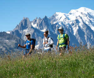 Tre vandrere går gennem en græsklædt eng med høje bjerge og sneklædte tinder i baggrunden. De er iført rygsække og afslappet vandretøj på en klar, solrig dag.