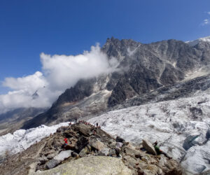 Landskabsudsigt over en gletsjer med forrevne bjerge i baggrunden. En gruppe vandrere ses på stenet terræn nær gletsjeren. Himlen er klar med nogle skyer omkring bjergtoppene.