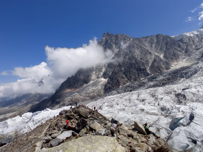 Landskabsudsigt over en gletsjer med forrevne bjerge i baggrunden. En gruppe vandrere ses på stenet terræn nær gletsjeren. Himlen er klar med nogle skyer omkring bjergtoppene.