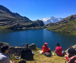 En gruppe mennesker sidder på en græsklædt bred og nyder en picnic ved en fredfyldt bjergsø. Sneklædte tinder og forrevne bakker danner en fantastisk kulisse under en klar blå himmel.