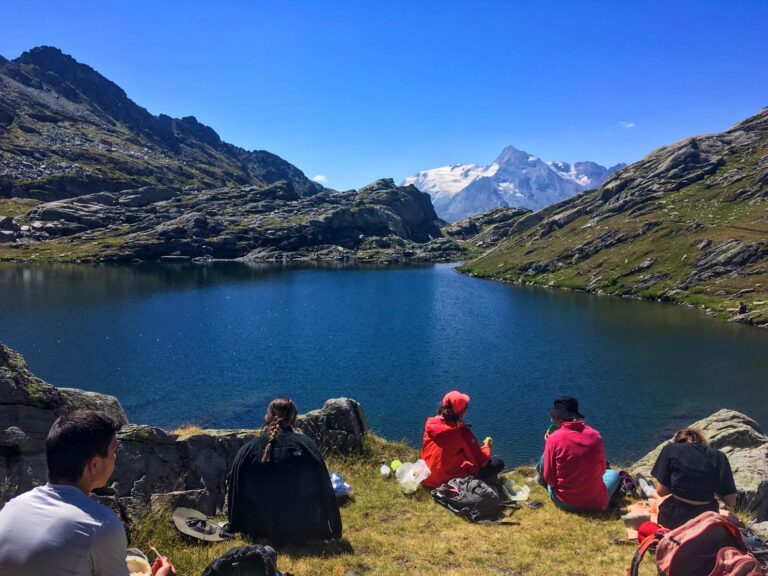 En gruppe mennesker sidder på en græsklædt bred og nyder en picnic ved en fredfyldt bjergsø. Sneklædte tinder og forrevne bakker danner en fantastisk kulisse under en klar blå himmel.