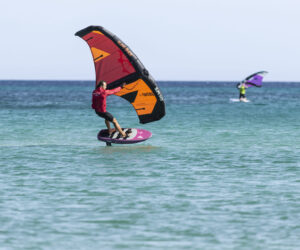 En person i en rød skjorte vingefoiling på et blåt og orange vingebræt over klart turkisblåt vand. En anden wingfoiler er synlig i baggrunden med en lilla vinge. Himlen er klar og havet er roligt.