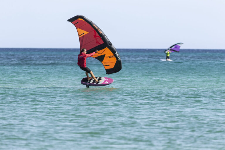 En person i en rød skjorte vingefoiling på et blåt og orange vingebræt over klart turkisblåt vand. En anden wingfoiler er synlig i baggrunden med en lilla vinge. Himlen er klar og havet er roligt.