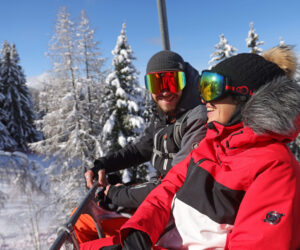 To personer i vinterudstyr sidder på en skilift omgivet af snedækkede træer. Begge er smilende og iført beskyttelsesbriller og varme jakker, med en klar blå himmel i baggrunden.