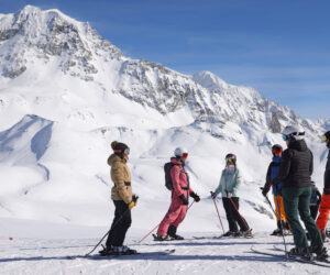 En gruppe skiløbere står på en sneklædt bjergskråning, omgivet af snedækkede tinder under en klar blå himmel. De er iført vinterudstyr og ser ud til at tale med skistave i hånden.