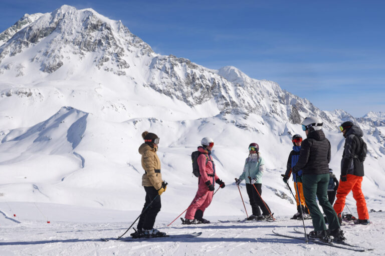 En gruppe skiløbere står på en sneklædt bjergskråning, omgivet af snedækkede tinder under en klar blå himmel. De er iført vinterudstyr og ser ud til at tale med skistave i hånden.
