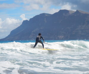 En surfer i en våddragt rider på et gult surfbræt på en lille bølge i havet. Himlen er overskyet, og en stor, barsk bjergkæde er synlig i baggrunden.
