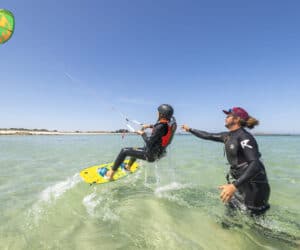 En kiteboarder i hjelm og redningsvest kører på et gult bræt på klart, lavt vand. En person i en våddragt står ved siden af dem og tilbyder vejledning. En farverig drage flyver højt mod en klar blå himmel. Hvid sandstrand i baggrunden.