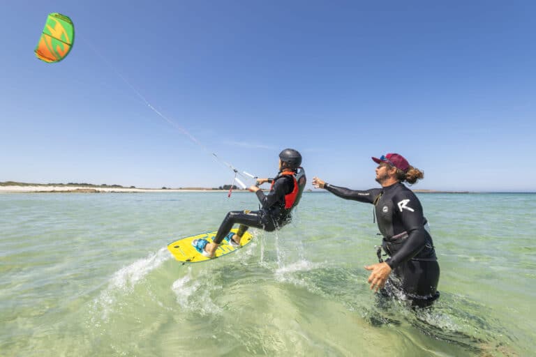 En kiteboarder i hjelm og redningsvest kører på et gult bræt på klart, lavt vand. En person i en våddragt står ved siden af dem og tilbyder vejledning. En farverig drage flyver højt mod en klar blå himmel. Hvid sandstrand i baggrunden.