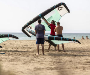 Tre mænd er på en sandstrand med store wingfoil. To brædder er turkise og sorte. Havet er synligt i baggrunden, og nogle få mennesker er i vandet. Himlen er klar.