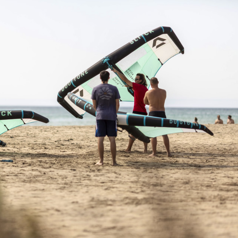 Tre mænd er på en sandstrand med store wingfoil. To brædder er turkise og sorte. Havet er synligt i baggrunden, og nogle få mennesker er i vandet. Himlen er klar.