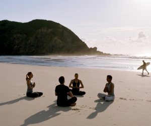 Fire personer sidder overskrævs i en meditationscirkel på en sandstrand, over for hinanden. Solen står lavt og skaber lange skygger. I baggrunden går en person med et surfbræt langs kysten nær klippebakker.