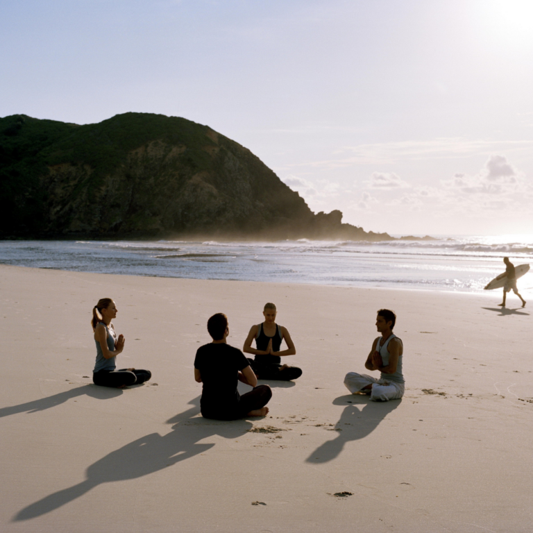 Fire personer sidder overskrævs i en meditationscirkel på en sandstrand, over for hinanden. Solen står lavt og skaber lange skygger. I baggrunden går en person med et surfbræt langs kysten nær klippebakker.