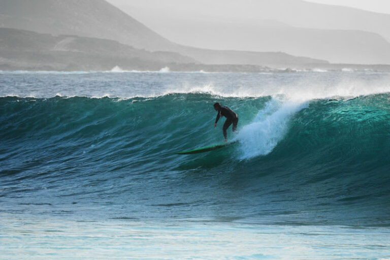 En person i en våddragt, der surfer på en stor havbølge under en overskyet himmel. Fjerne bakker er synlige i baggrunden, hvilket tilføjer dybde til scenen.